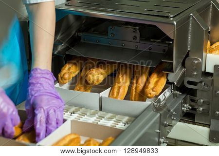 Boxes of eclairs on conveyor. Hands working with baked pastry. Machine puts eclairs into boxes. Each worker has a task.