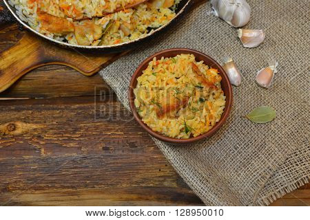 Vintage clay bowl with traditional spicy food pilaf, frying pan with pilaf on wooden background and the burlap. Pilaf with meat