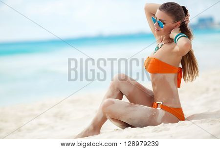 Young beautiful brunette woman with long straight hair,mirrored sun glasses,blue, slim figure,wearing around his neck a large necklace,on the left arm-bracelets, spends time on a tropical beach on the white sands in an orange bikini near blue ocean