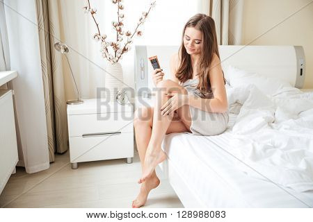 Happy young woman putting cream on legs on the bed at home