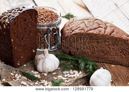 Close up photo of rustic bread sprinkled with sunflower seeds on sackcloth, garlic, dill, oat flakes and jar with buckwheat on planked wooden table