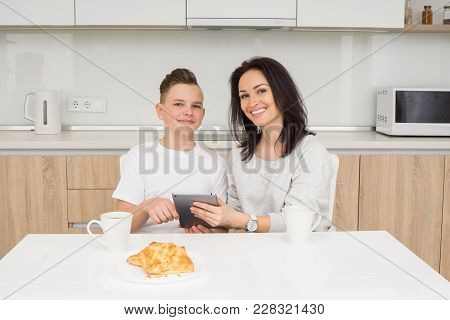 Mother And Son Using Digital Tablet Together Sitting At Table In Kitchen. Smiling And Looking At Cam