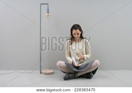Beautiful Girl Reads A Book While Sits On The Floor In Lotus Pose Next To The Long Lamp With Edison 
