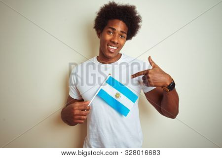 Afro american tourist man holding Argentinian Argentina flag over isolated white background with surprise face pointing finger to himself