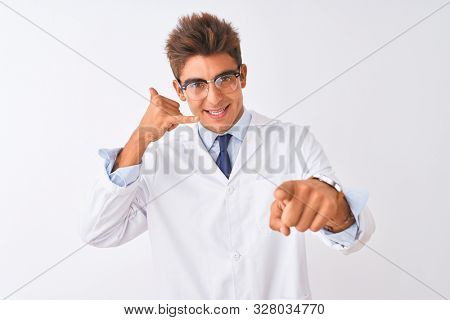 Young handsome sciencist man wearing glasses and coat over isolated white background smiling doing talking on the telephone gesture and pointing to you. Call me.