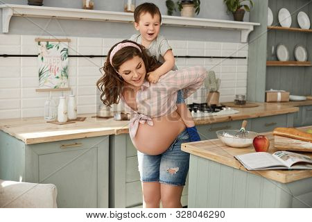 A Happy Young Pregnant Woman Has Fun With Her Little Son And Making Breakfast At The Kitchen In The 