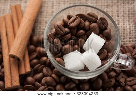 Coffee Beans And Sugar In Glass Cup On Sackcloth With Cinnamon Sticks Beside. Scattered Coffee Beans