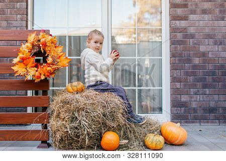 Adorable Smiling Caucasian Baby Toddler In White Knittes Jacket Sitting On The Haystack With Pumpkin