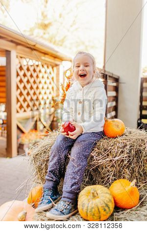 Adorable Blonde Baby Toddler In White Knittes Jacket Sitting On The Haystack With Pumpkins At Porch,