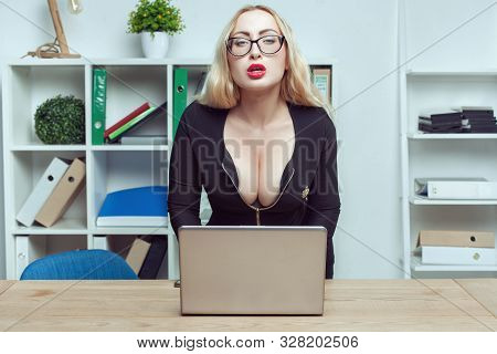 Young Woman Secretary Sits At A Table In The Office. She Works Behind A Laptop And Looks In Front Of