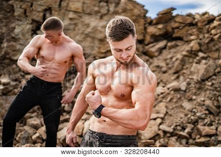 Two Fitness Strongmen Pumping Muscles In A Rocky Background. Bodybuilders Concept Background - Muscu