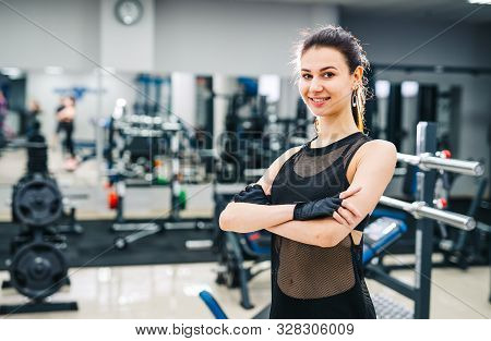Young Beautiful Sportwoman With Nice Body. Brunette Fitness Girl With Crossed Hands. Gym Background.