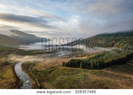 Sun Is Setting As Low Hanging Clouds Run Through The River Orchy, A Pine Forest And The Hills In The