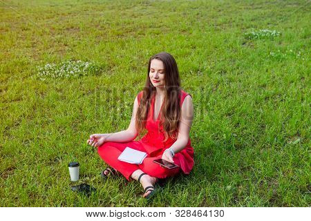 Young Businesswoman In Red Suit Sitting In Lotus Position On Green Grass In Park And Holding Her Sma
