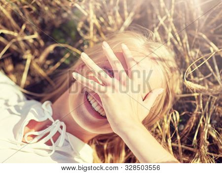 nature, summer holidays, vacation and people concept - close up of happy young woman lying on cereal field and covering face by hand