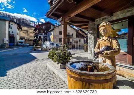Livigno, Italy, April 2016 Traditional Alpine Wooden Architecture In The Village, Fountain And Sculp