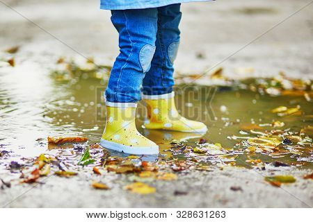Child Wearing Yellow Rain Boots And Jumping In Puddle On A Fall Day. Toddler Girl Having Fun With Wa