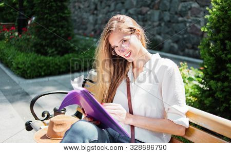 Young Caucasian Woman Student In Casual Clothing Reading Esse In Folder While Sitting On Bench At Pa