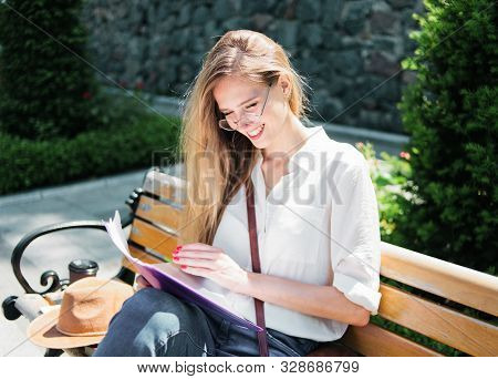 Young Caucasian Woman Student In Casual Clothing Reading Esse In Folder While Sitting On Bench At Pa