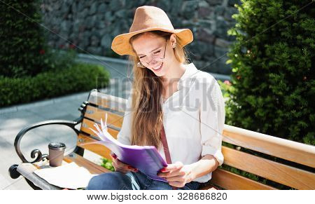 Young Caucasian Woman Student In Casual Clothing Reading Esse In Folder While Sitting On Bench At Pa