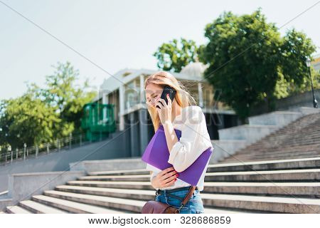 Young Caucasian Woman Student In Casual Clothing With Esse In Folder Talking On Phone Going Down The