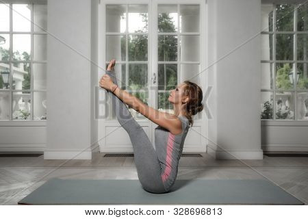 Beautiful young woman sitting in Paripurna Navasana pose, practicing meditation in  hall 