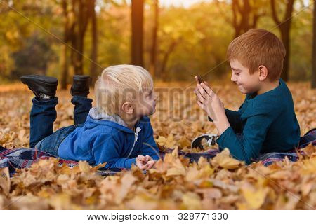 Two Little Brothers Take Pictures Of Each Other, Lying In Yellow Autumn Leaves. Fall Day
