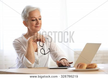 Mature Woman With Laptop Sitting At Table In Kitchen. Smart Aging