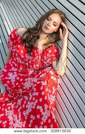 Beautiful Young Girl, Model, Brown-haired Woman With Long Hair In A Red Dress Sits On A Bench And Po
