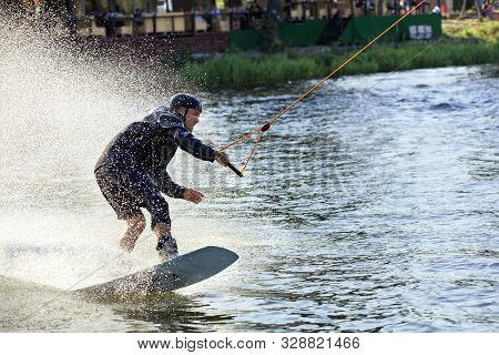 Wakeboarder Rushes Through The Water Along The Grassy Banks Of The River, Holding The Cable With One