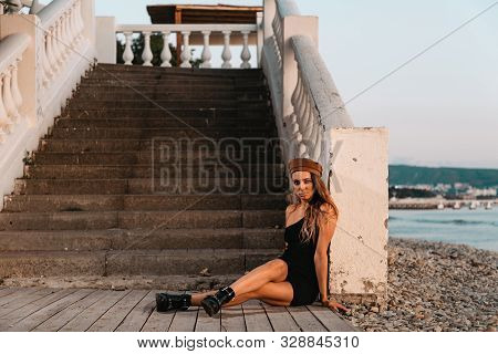 Fashion. Sexy Blonde In A Black Dress And Hat Posing Sitting On The Steps. In The Distance You Can S