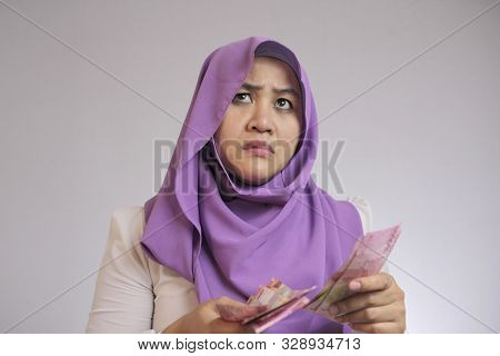 Confused Stressed Asian Muslim Woman Holding Indonesian Rupiah Cash In Hand, Thinking Wondering How 