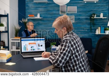 Elderly Senior Woman Taking Notes In Notebook While Working On Laptop In Cozy Living Room. Adult Int