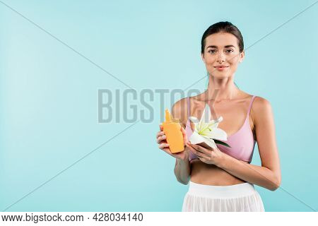 Young Woman With Bottle Of Sunscreen And White Lily Looking At Camera Isolated On Blue