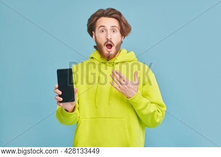 Funny young man in light green hoddie is holding the phone in hi hands and is very surprising. Studio portrait on a blue background with copy space.