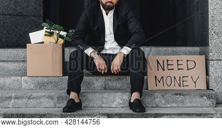 Hopeless Businessman With Paper Poster Saying Need Money And Box Of Office Stuff, Sitting On Stairs,