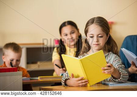 Girl Reading Book During Leson In Montessori School Near Blurred Multiethnic Friends