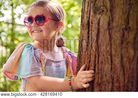 Smiling Young Girl Wearing Sunglasses Playing Hide And Seek Behind Tree In Garden