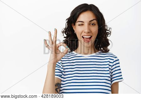 Cheerful Curly Girl With Pierced Nose, Winking And Showing Ok Okay Gesture, No Proble, Have Things U