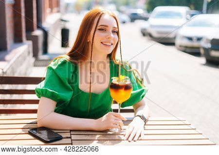 Close-up Portrait Of Happy Smiling Young Woman Drinking Cocktail Through Straw Sitting At Table In O