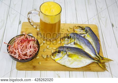 A Mug Of Beer With Foam, Dried Vomer Fish And Squid Shavings In A Round Black Plate On A Wooden Boar