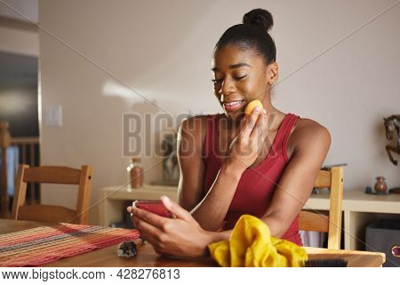 african american woman applying makeup with spronge and using smartphone as mirror