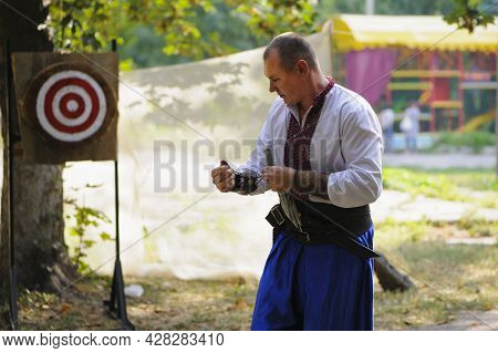 A Man Throws Knives At A Wooded Target. White Shirt And Blue Pants On A Young Guy. National Clothes.