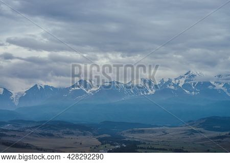 Scenic Mountain Landscape With Great Snowy Mountain Range Among Low Clouds And Green Forest In Valle