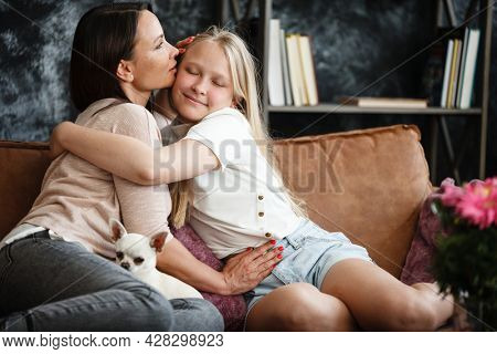 Mother and daughter relationship concept. A woman and a teenage girl are sitting in front of the TV on the couch and happy embarcing. The family spends time at home. Little dog lying on laps.