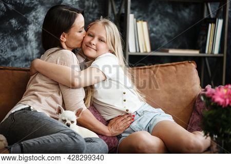 Mother and daughter relationship concept. A woman and a teenage girl are sitting in front of the TV on the couch and happy embarcing. The family spends time at home. Little dog lying on laps.