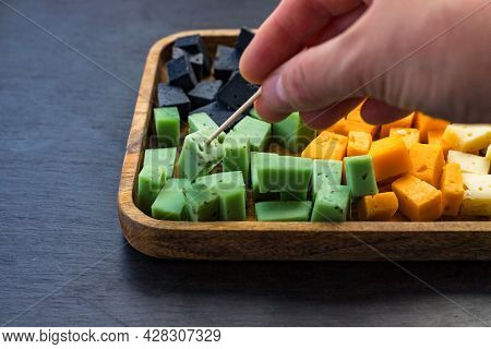 Human Hand Pricking Piece Of Cheese Multycolored Cubes On Wooden Tray On Black Slate Background. Che