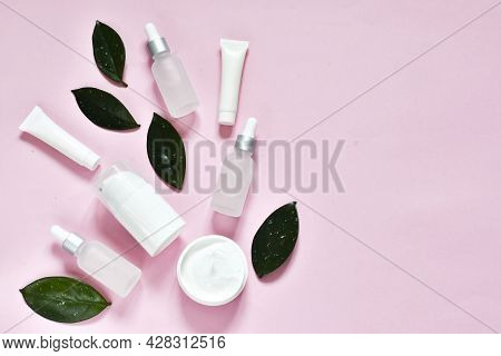 White Glass Cosmetic Bottles Pipette And Jars With Green Leavers On A Pink Background. Top View. Spa