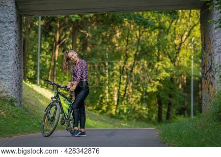 Young Happy Woman In Leggings And A Shirt With A Bicycle Stands On An Asphalt Road In A Park Under A