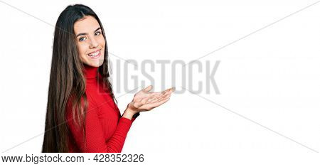Young brunette teenager wearing red turtleneck sweater pointing aside with hands open palms showing copy space, presenting advertisement smiling excited happy 